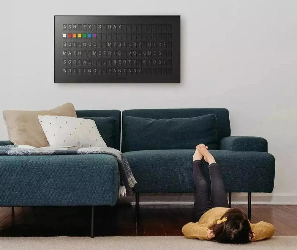 Black Vestaboard split-flap display above a teal sofa in a bright living room