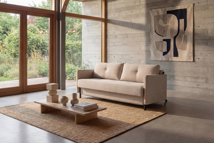 Cubed sofa bed with arms in beige fabric in a sunlit room with floor-to-ceiling windows