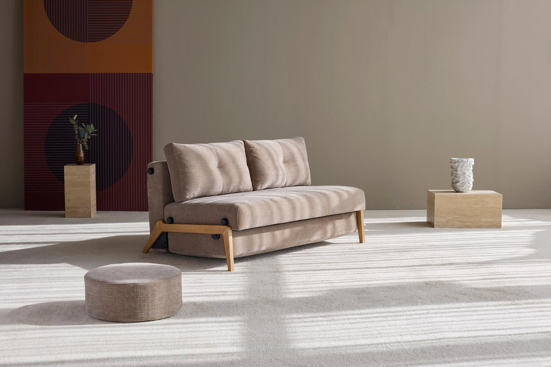 Cubed sleeper sofa with oak legs in taupe fabric on a light rug with a pouf