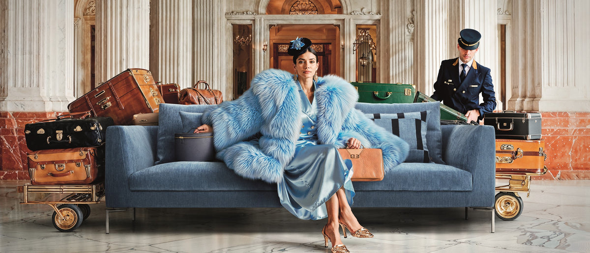 Blue Eilersen Ra sofa in a grand hotel lobby with marble columns and vintage luggage.