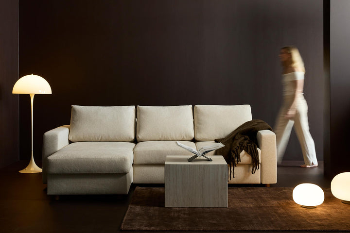 Neah Sleeper Chaise Sectional by Innovation Living in cream with chaise as a woman walks past a globe floor lamp